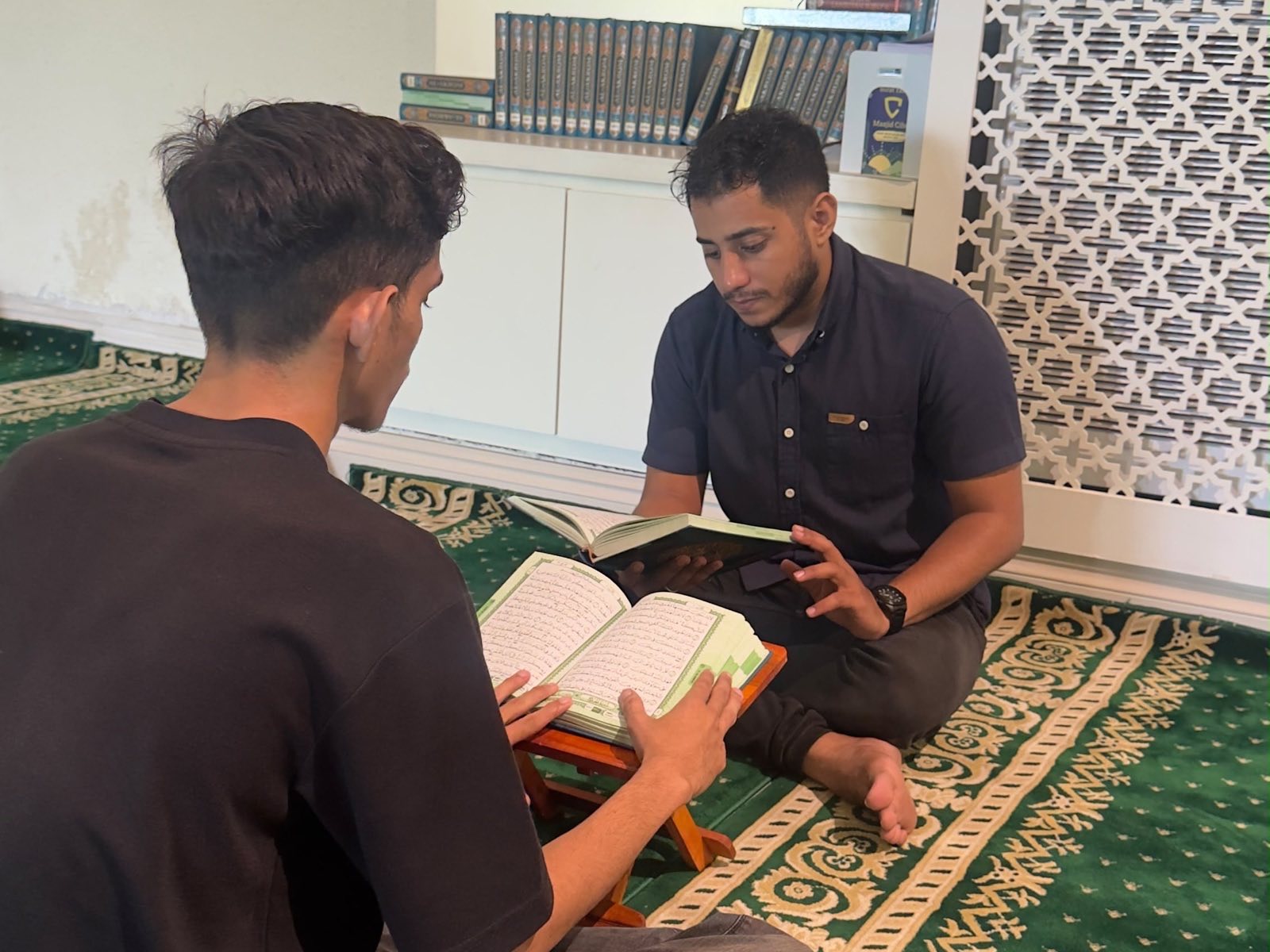 Seorang guru berbatik dan satu murid duduk berhadapan, rehal berisi mushaf di antara keduanya